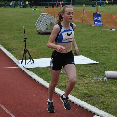 Laufende Sportlerin in Wettkampfoutfit auf einer roten Laufbahn mit Zuschauern im Hintergrund.