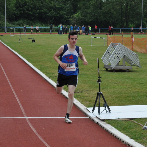 Ein junger Läufer sprintet auf einer roten Tartanbahn beim Wettkampf, im Hintergrund sind Zuschauer und Sportgeräte sichtbar.