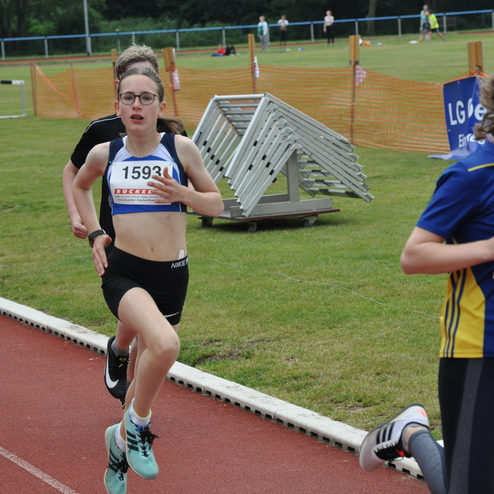 Junge Athletin läuft auf der Bahn, trägt Sportkleidung mit Startnummer 1593, im Hintergrund andere Sportler und Hindernisse.