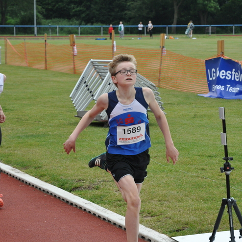 Junger Athlet mit der Startnummer 1589 sprintet auf einer Laufbahn, im Hintergrund sind andere Sportler und eine Wettkampfkulisse.