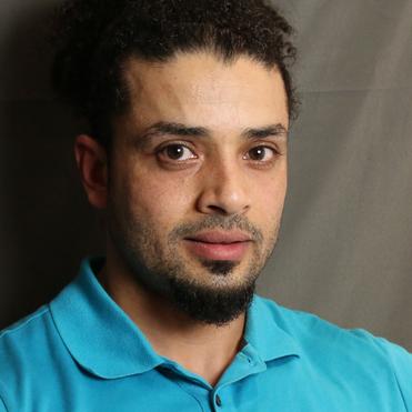 Nahaufnahme eines Mannes mit lockigem Haar, getragenem blauen Poloshirt, vor neutralem Hintergrund.