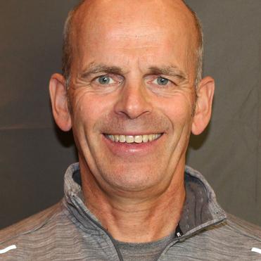 Lächelnder Mann in grauem Sportpullover mit Reißverschluss, vor einem grauen Hintergrund.