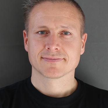 Porträt eines Mannes mit kurzem, gestyltem Haar, in schwarzem T-Shirt, vor neutralem grauem Hintergrund.