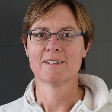 Portrait einer Frau mit kurzen, blonden Haaren und Brille, die in einem weißen Hoodie vor einem grauen Hintergrund steht.