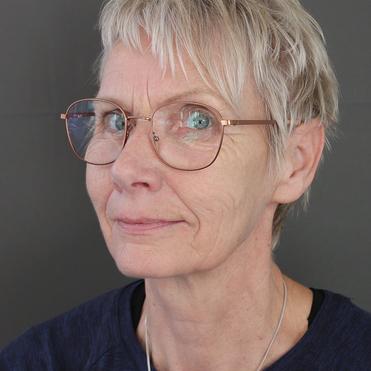 Nahaufnahme einer Frau mit kurzen, blonden Haaren und modischen Brillen, neutralem Gesichtsausdruck vor grauem Hintergrund.