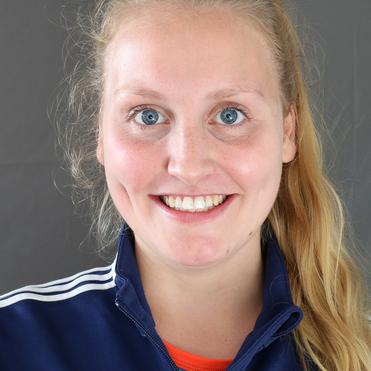 Porträt einer lächelnden Frau mit blonden Haaren, blauen Augen und Sportjacke vor einem grauen Hintergrund.