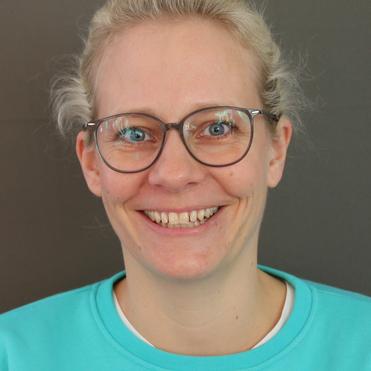 Porträt einer lächelnden Frau mit Brille, blonden Haaren, in einem türkisfarbenen Sweatshirt, vor grauem Hintergrund.