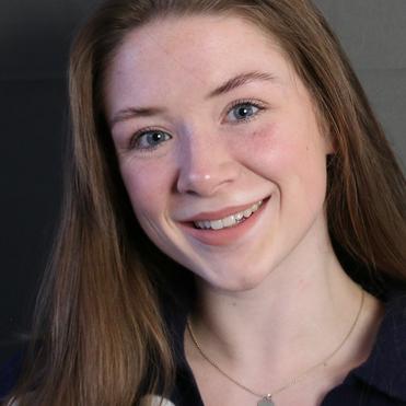 Porträt einer jungen Frau mit langen, hellbraunen Haaren, lächelnd, in einem dunklen Oberteil und mit einer Halskette.