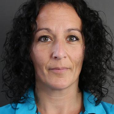 Porträt einer Frau mit lockigem Haar, die ein blaues Poloshirt trägt, vor einem grauen Hintergrund.