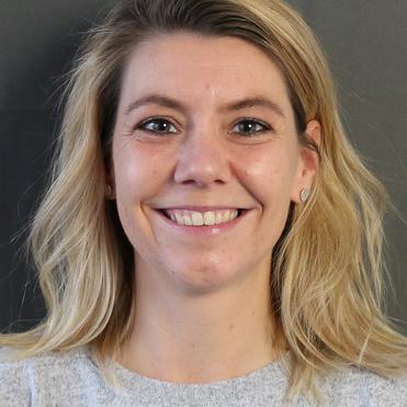 Porträt einer lächelnden Frau mit blonden, lockigen Haaren, die ein graues Sweatshirt trägt, vor einem grauen Hintergrund.