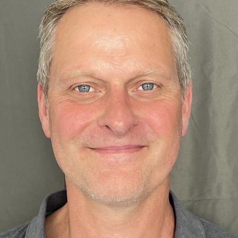 Porträt eines lächelnden Mannes mit grauen Haaren und blauen Augen, vor einem grauen Hintergrund.