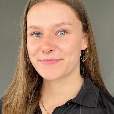 Nahaufnahme einer lächelnden jungen Frau mit langen, glatten Haaren, in schwarzem Poloshirt vor grauem Hintergrund.