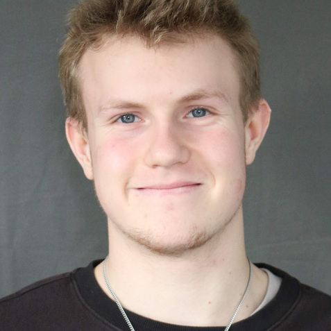 Portrait eines lächelnenden jungen Mannes mit kurzen, blonden Haaren, im schwarzen Pullover vor grauem Hintergrund.