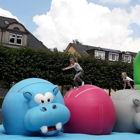 Zwei Kinder springen auf großen, farbigen, hüpfenden Ball-Tieren auf einem Spielplatz unter einem blauen Himmel.