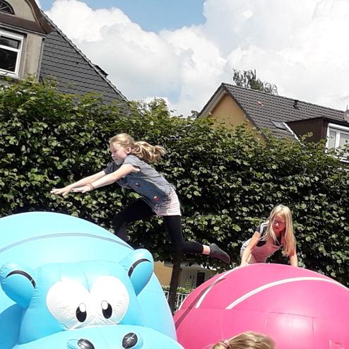 Zwei Kinder springen auf großen, bunten Hüpfbällen in einem Freiluftbereich, umgeben von grünen Büschen und Häusern.