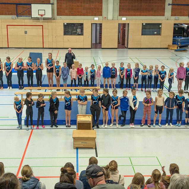 Gruppe von Gymnastinnen in blauen Trikots versammelt in einer Sporthalle, mit Zuschauern im Hintergrund.