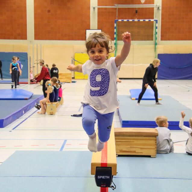 Ein kleines Kind mit Shirt in Weiß und lila auf einem Sprungbrett, während andere Kinder im Sportbereich trainieren.