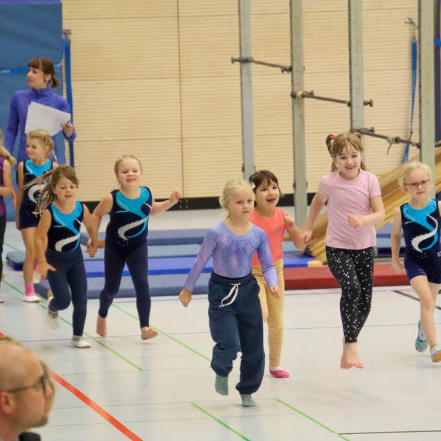Gruppe von Kindern in Sportanzügen, die fröhlich in einer Turnhalle laufen, während Betreuer sie beobachtet.