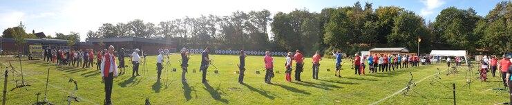 Panoramablick auf Schützen in Sportkleidung beim Bogenschießen auf einem grünen Feld mit Zielscheiben im Hintergrund.