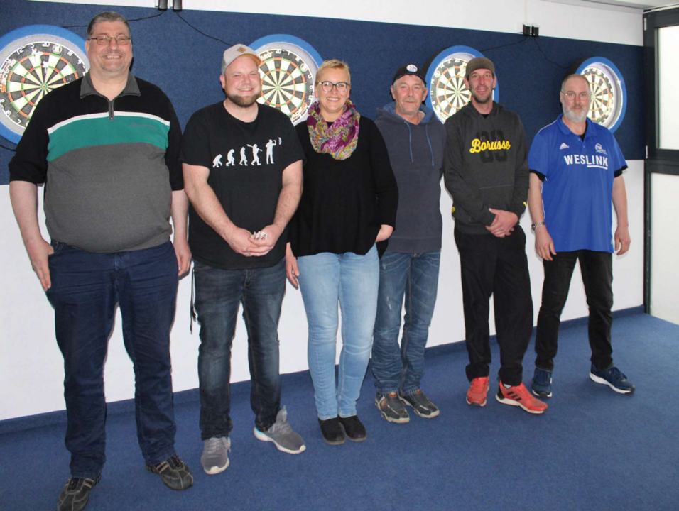 Sechs Personen stehen in einer Dart-Spielhalle vor zwei Dartboards an der Wand.