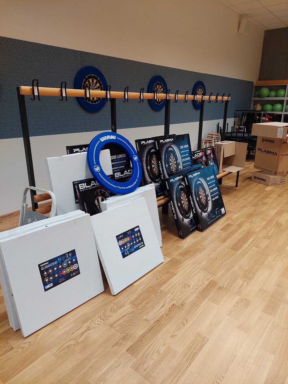 Dartboards und Zubehör sind in einem Raum auf einem Holzfußboden organisiert, mit gestapelten Platten und Kisten.