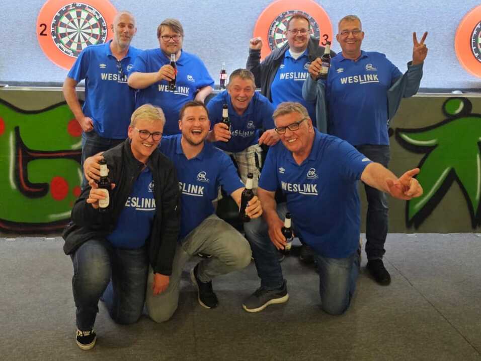 Gruppe von acht Männern in blauen „WESLINK“-Shirts posiert lächelnd mit Flaschen auf einem Darts-platz.