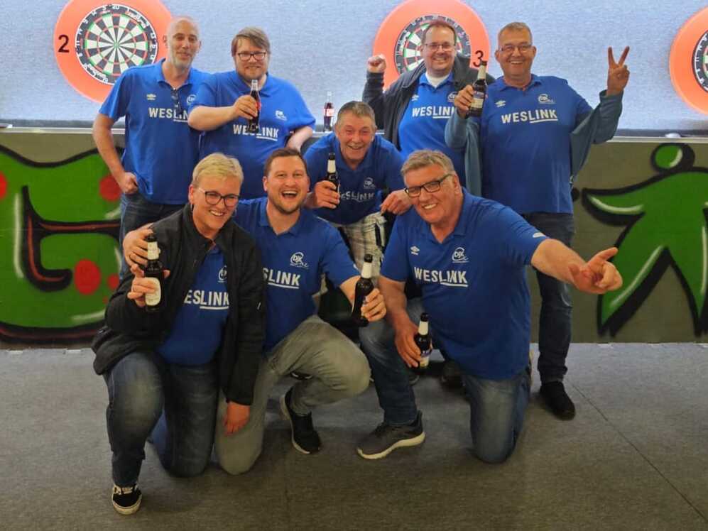Gruppe von acht Männern in blauen „WESLINK“-Shirts posiert lächelnd mit Flaschen auf einem Darts-platz.