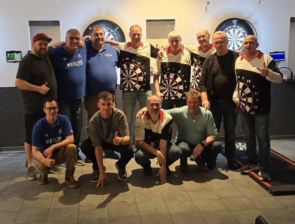 Gruppe von neun Männern in Darttrikots, posierend vor Dartboards in einem Spielraum. Einige zeigen Daumen hoch.