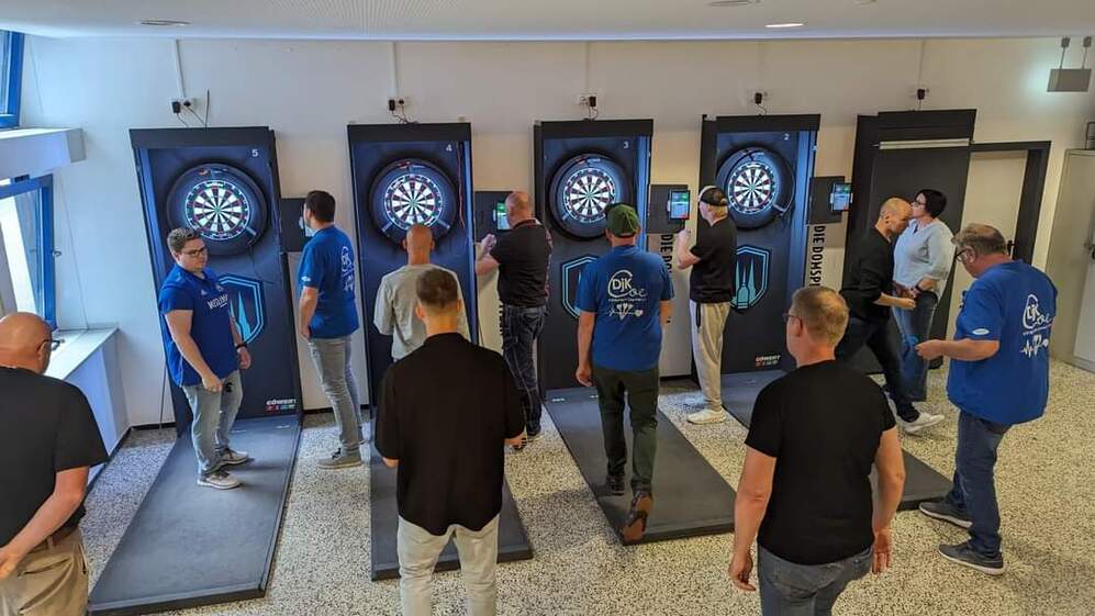 Gruppenspieler werfen Darts auf elektronischen Targetwänden in einem hellen, modernen Raum.