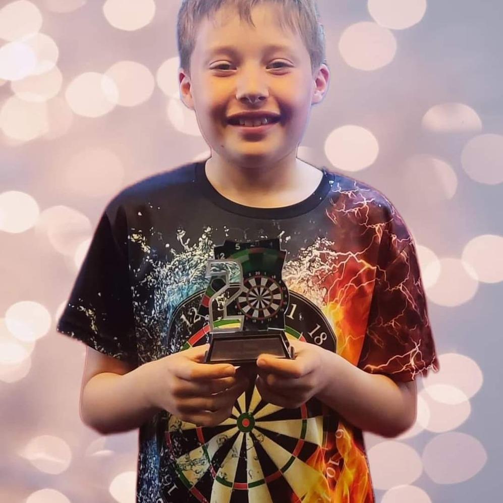 Lächelnder Junge in einem bunten T-Shirt hält eine kleine Dartscheibe vor einem unscharfen, glitzernden Hintergrund.