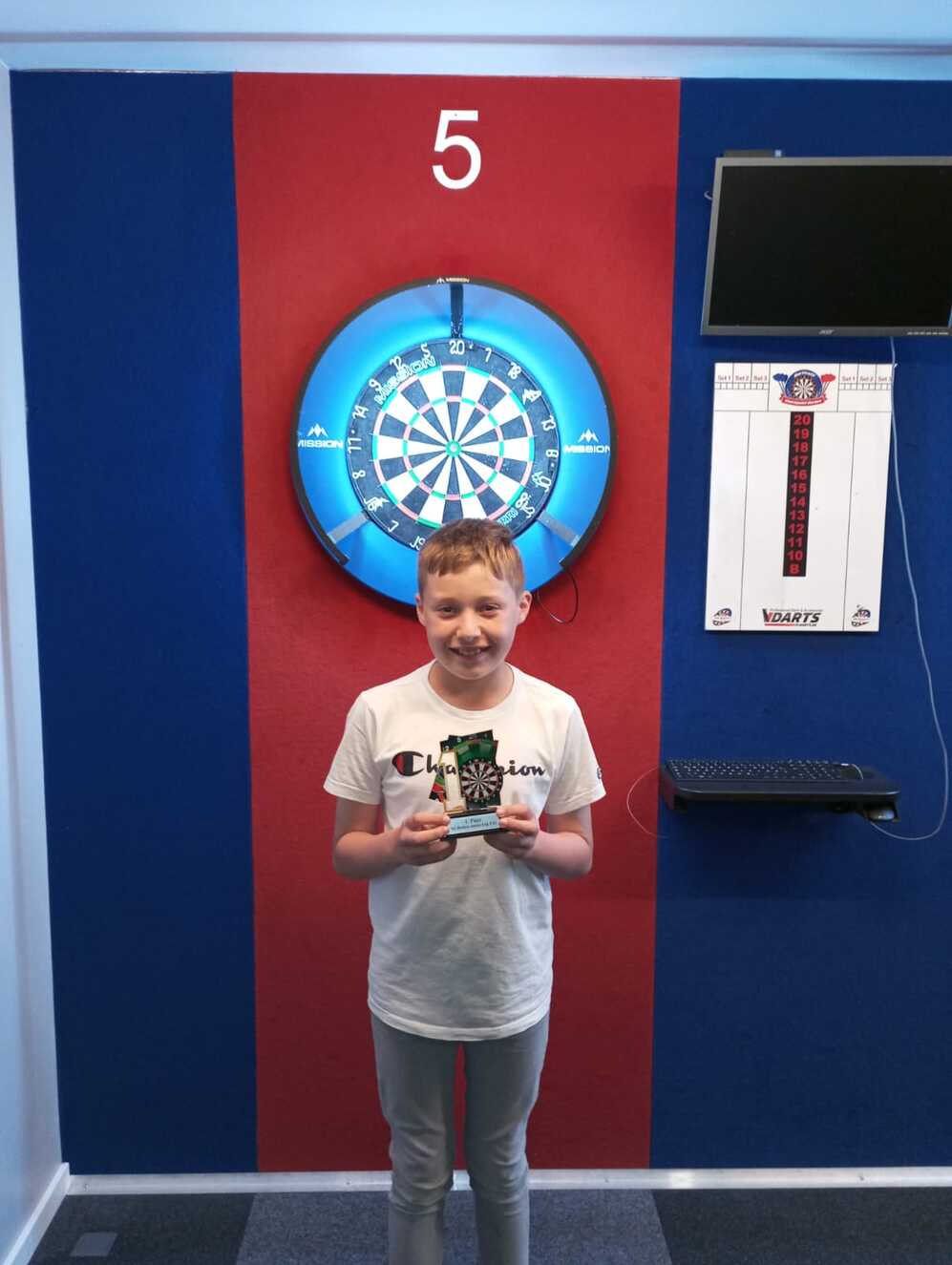 Junger Junge mit Pokal steht vor einer Dartscheibe in einem dartspielgerechten Raum.