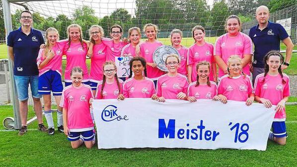 Mädchenfußballmannschaft in pinken Trikots posiert mit Meisterschaftsschild und Pokal auf dem Sportplatz.