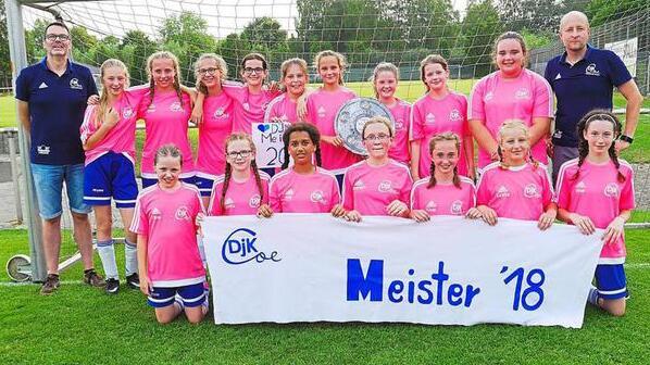 Mädchenfußballmannschaft in pinken Trikots posiert mit Meisterschaftsschild und Pokal auf dem Sportplatz.