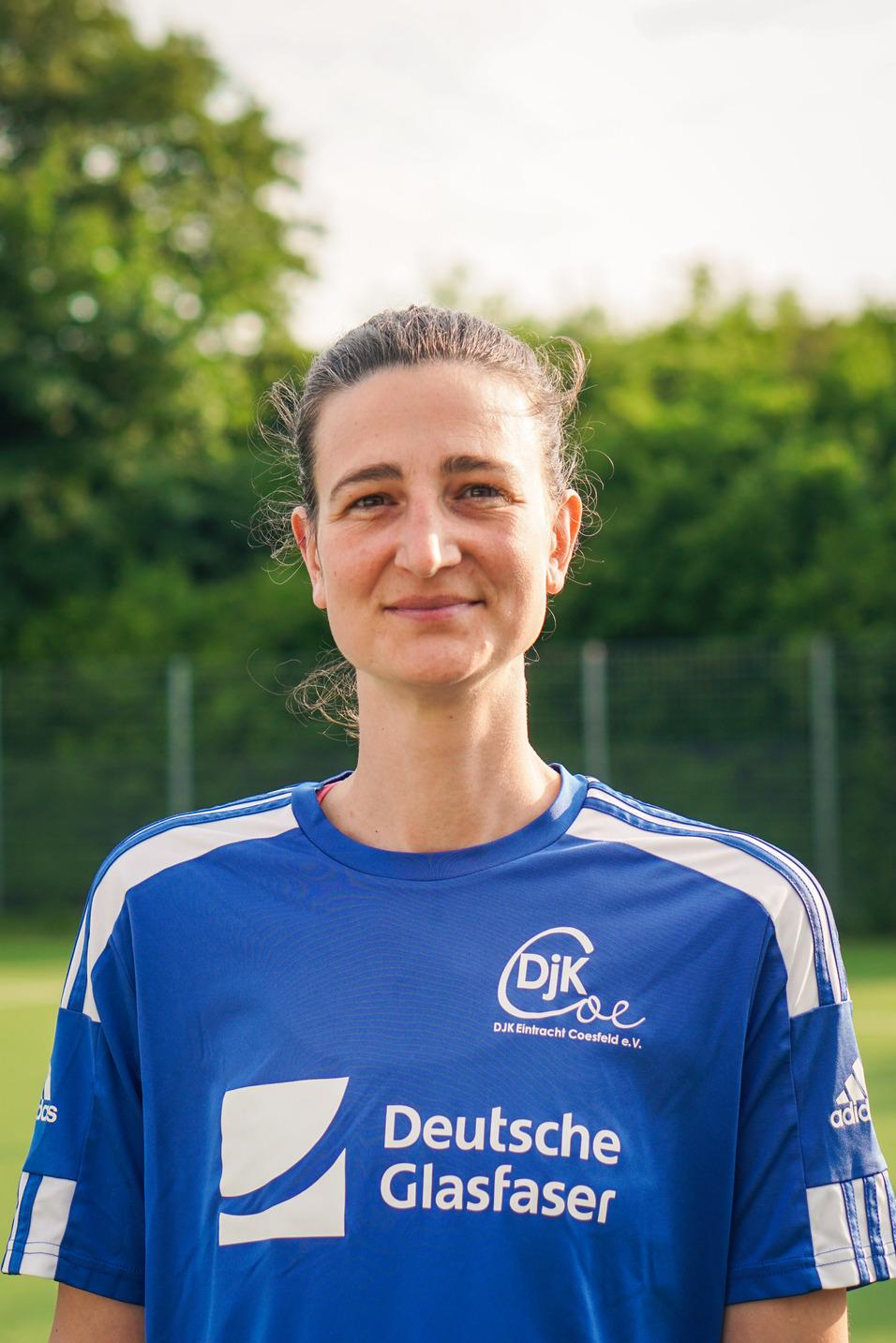 Porträt einer sportlichen Frau in einem blauen Trikot mit Vereinslogo, im Hintergrund ein grüner Sportplatz.