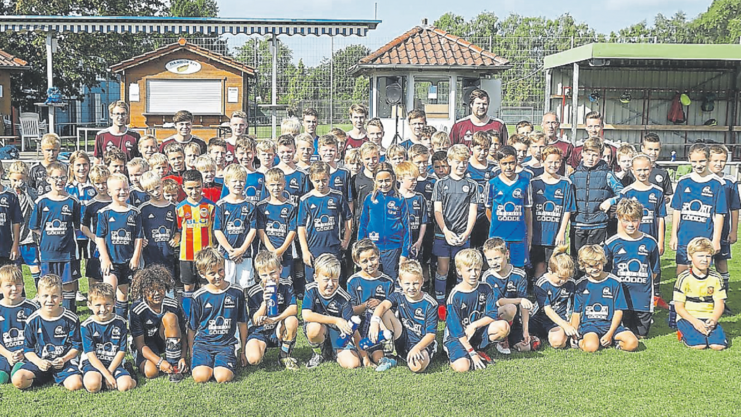 Gruppenfoto von Kindern in blauen Fußballtrikots auf einem Sportplatz, im Hintergrund Gebäude und Zuschauer.
