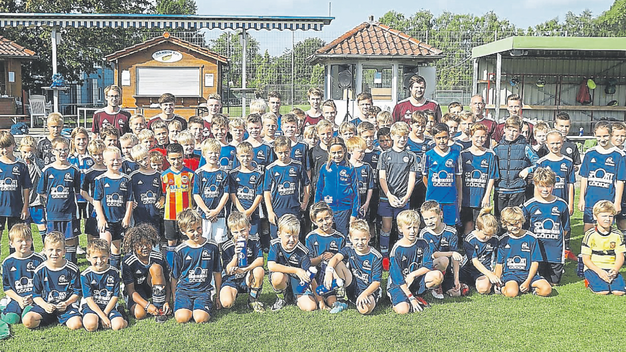 Gruppenfoto von Kindern in blauen Fußballtrikots auf einem Sportplatz, im Hintergrund Gebäude und Zuschauer.
