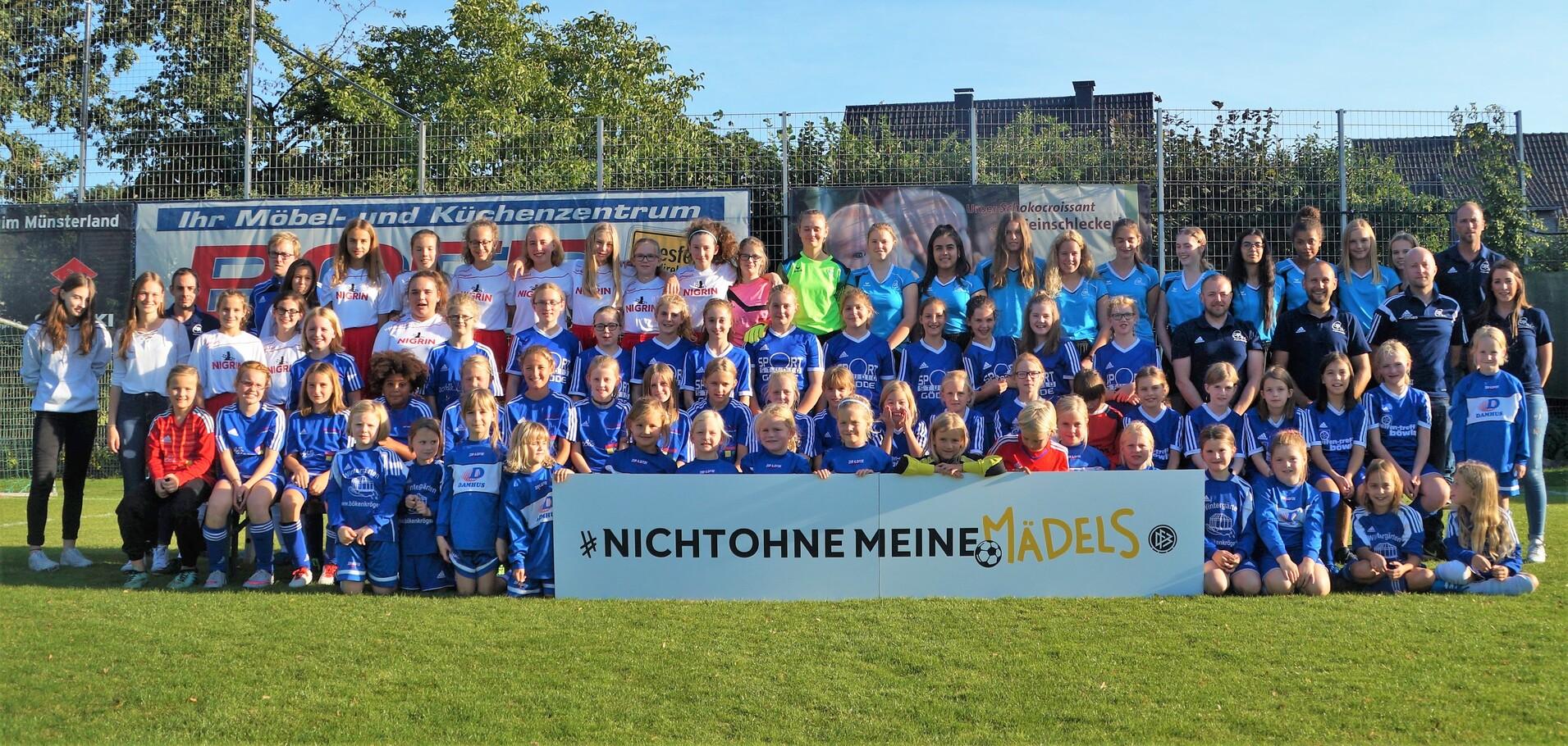 Gruppenfoto von Mädchen- und Frauenfußballspielern in blauen Trikots, posierend vor einem Banner mit dem Slogan „#NICHTOHNE MEINE MÄDELS“.