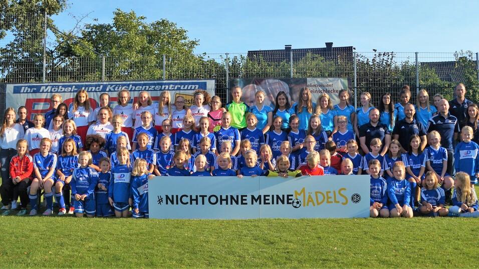 Gruppenfoto von Mädchen- und Frauenfußballspielern in blauen Trikots, posierend vor einem Banner mit dem Slogan „#NICHTOHNE MEINE MÄDELS“.