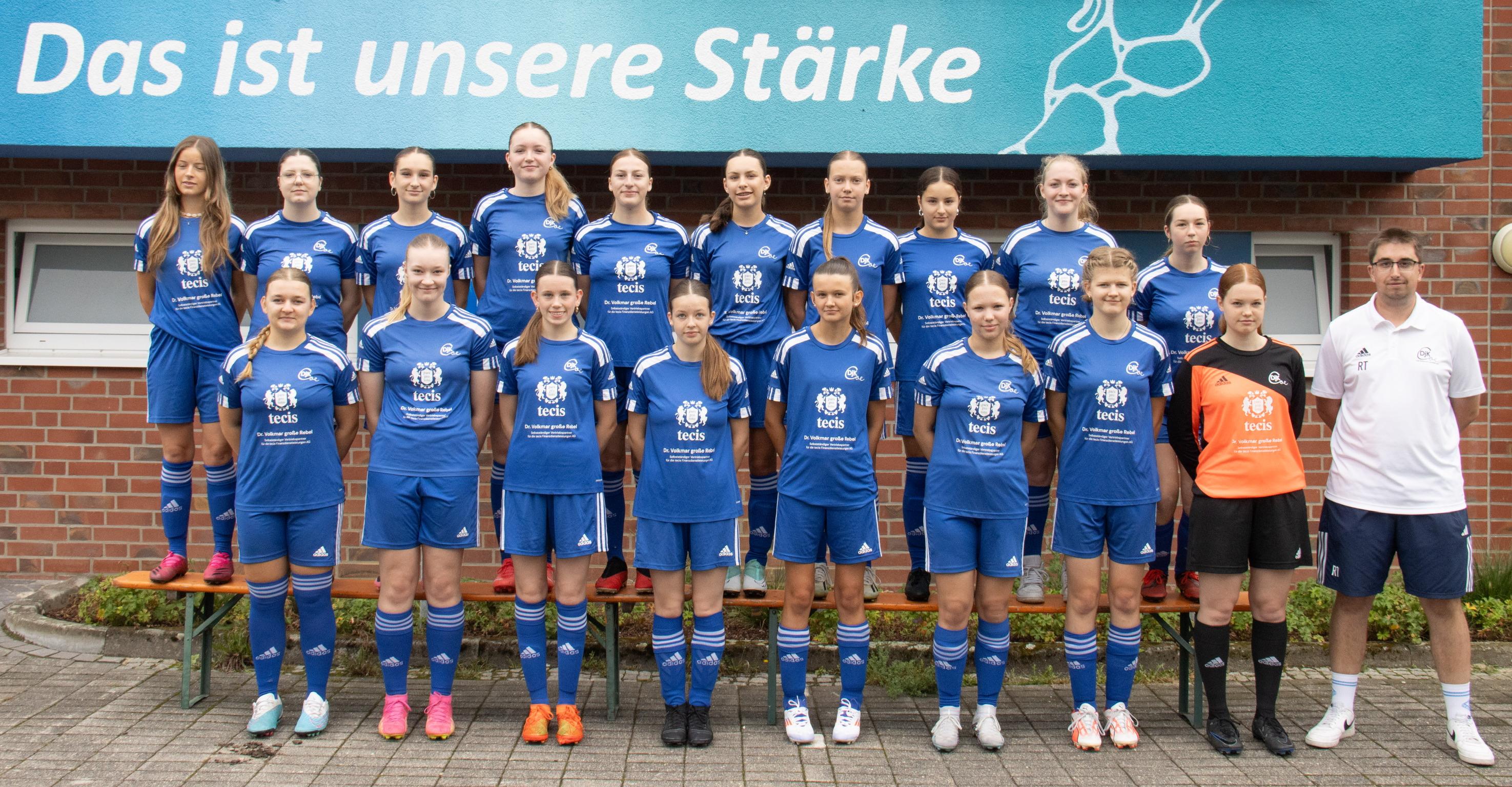 Mannschaftsfoto einer Frauenfußballmannschaft in blauen Trikots vor einer Wand mit dem Schriftzug „Das ist unsere Stärke“.