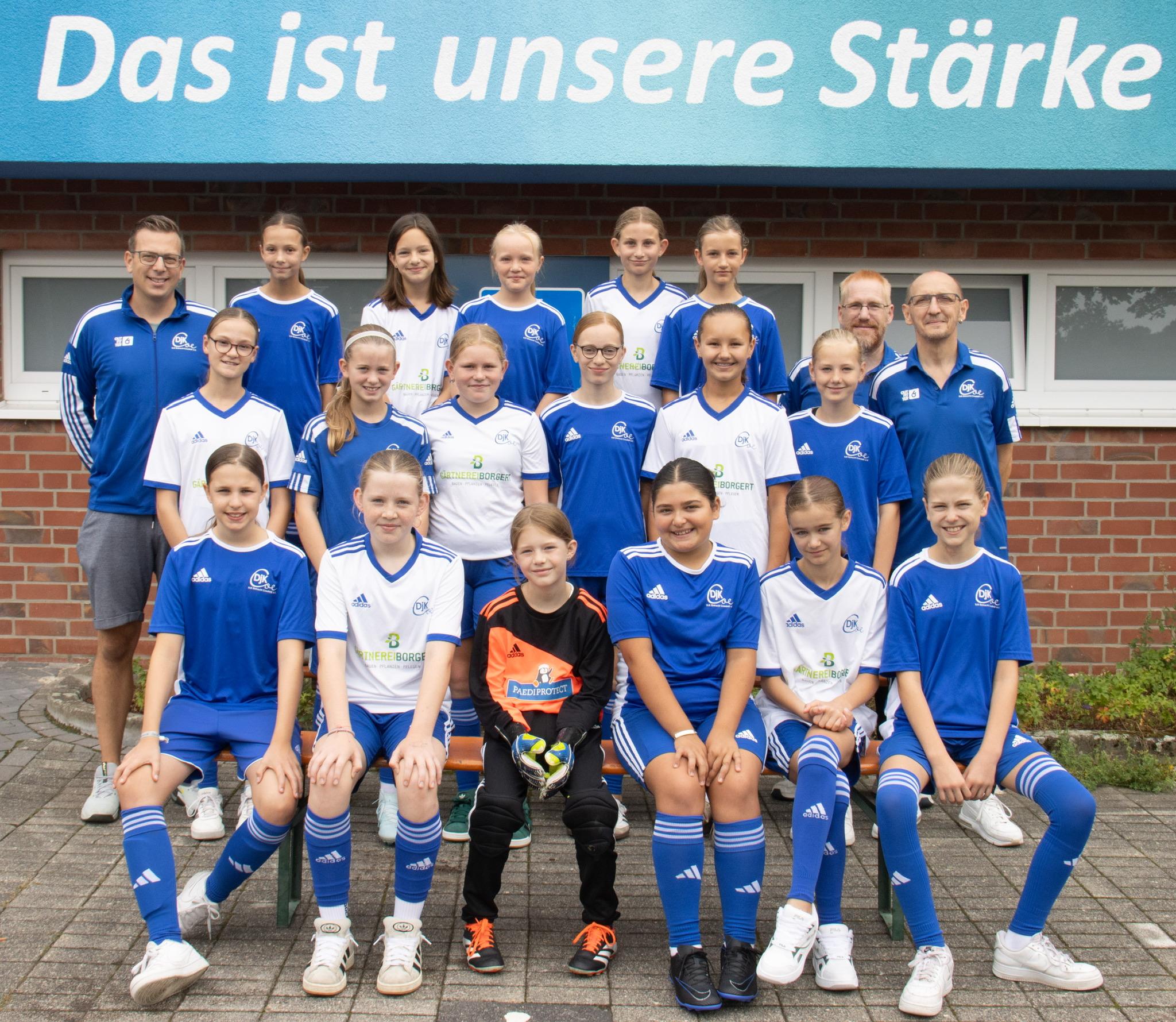Mannschaftsfoto einer Fußballjugend mit Trainer, alle in blauen und weißen Trikots, vor einem Gebäude mit Spruch.