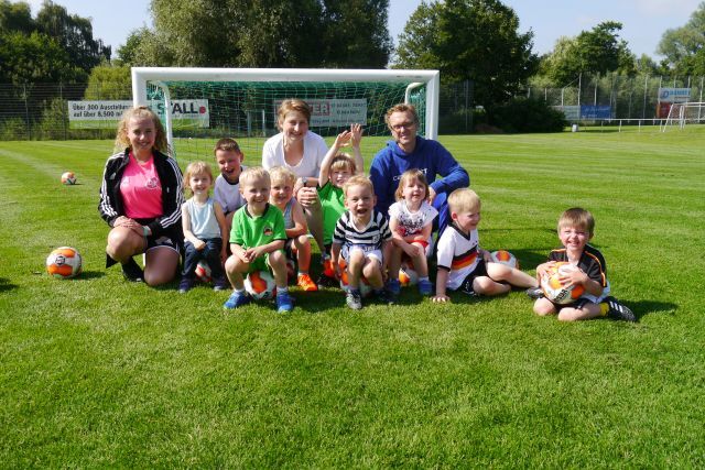 Gruppe von Kindern und Trainern, die lachend auf einem Fußballplatz vor einem Tor sitzen, umgeben von Fußbällen.