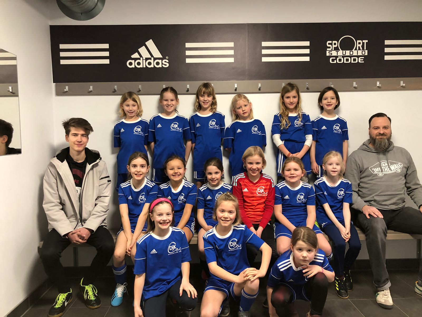 Mädchenfußballteam in blauen Trikots posiert vor einer Wand mit Adidas-Logo und Sportstudio Göddé-Schild.