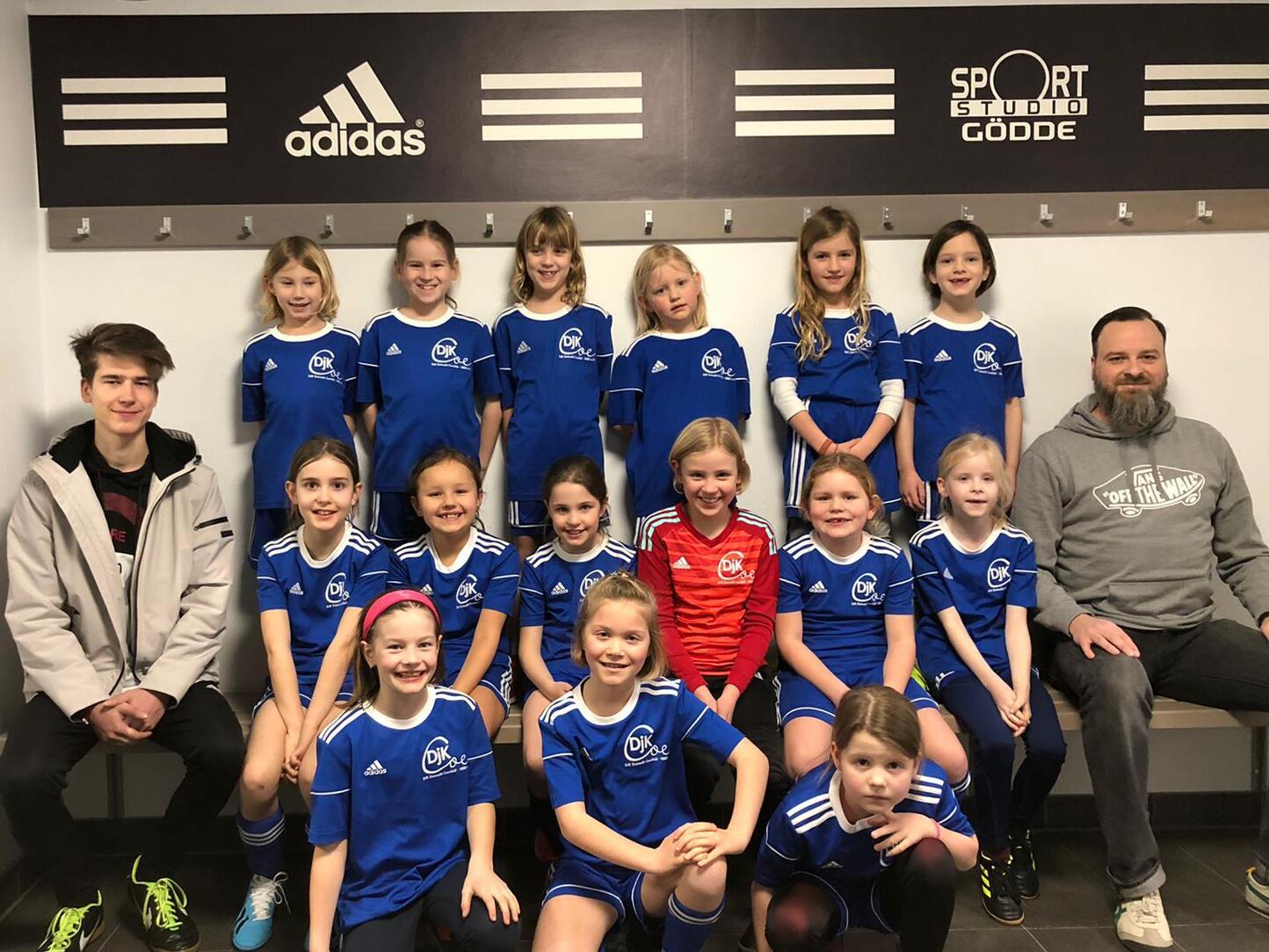M&auml;dchenfu&szlig;ballteam in blauen Trikots posiert vor einer Wand mit Adidas-Logo und Sportstudio G&ouml;dd&eacute;-Schild.