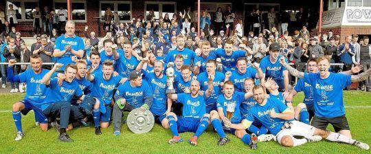 Feiernde Fußballmannschaft in blauen Trikots, posierend mit Pokal, umgeben von jubelnden Zuschauern auf einem Sportplatz.