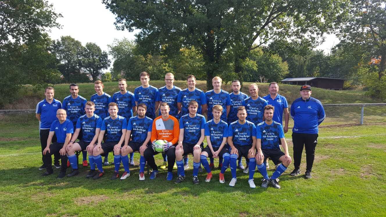 Gruppenfoto einer Fußballmannschaft in blauen Trikots auf einem Platz, umgeben von Bäumen und einem Gebäude im Hintergrund.