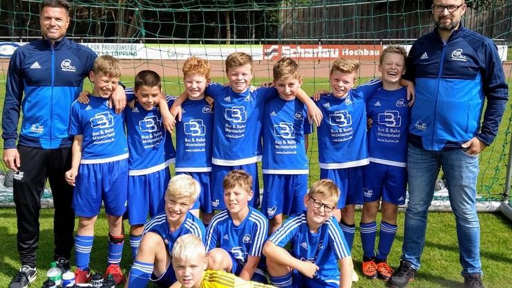 Gruppe von zw&ouml;lf Kindern im blauen Fu&szlig;balltrikot, posierend mit zwei Trainern auf einem Sportplatz.