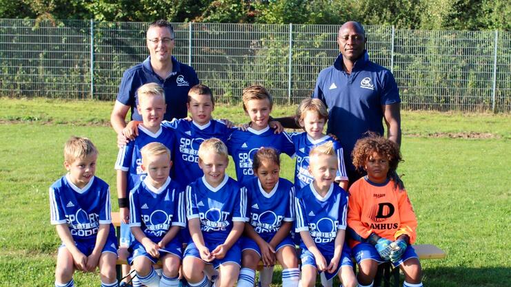 Gruppenfoto einer Jugendfußballmannschaft mit Trainern, alle in blauen Trikots, auf einem grünen Rasenplatz.