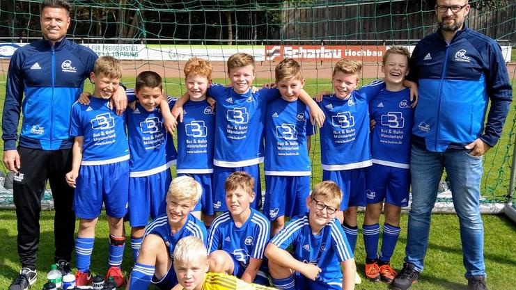 Gruppe von zwölf Kindern im blauen Fußballtrikot, posierend mit zwei Trainern auf einem Sportplatz.