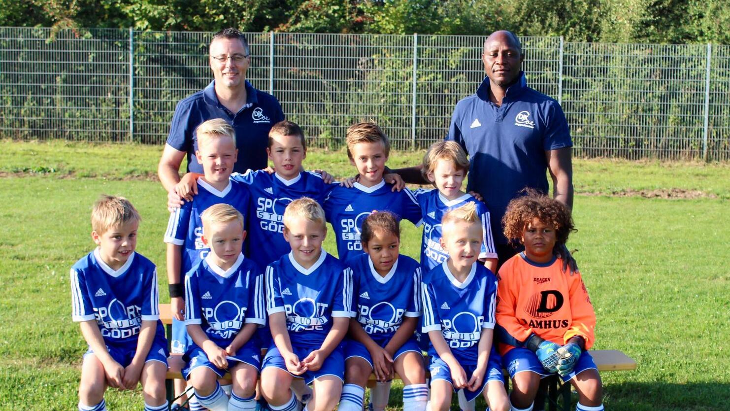 Gruppenfoto einer Jugendfußballmannschaft mit Trainern, alle in blauen Trikots, auf einem grünen Rasenplatz.