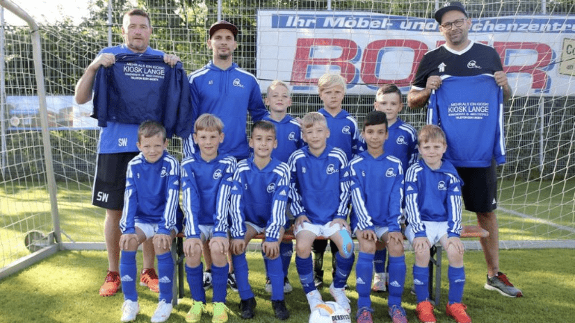 Eine Gruppe von Kindern in blauen Fußballtrikots posiert mit Trainern vor einem Tor und einem Banner.